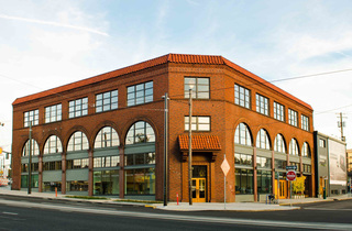An exterior image of a brick building with an entrance facing the corner and a red tile roof.